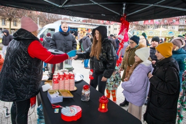 Stoisko na zewnątrz pod czarnym namiotem, kilka osób w zimowych ubraniach stoi w kolejce po napoje w papierowych kubkach z czerwonym świątecznym wzorem. Kobieta w różowej czapce i kamizelce podaje napój młodej kobiecie w czarnej kurtce z kapturem. Na stole są dekoracje świąteczne, w tym czerwony lampion i pudełko z Mikołajem. W tle widać więcej ludzi oraz budynki.