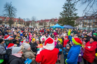 Duża grupa dzieci i dorosłych zgromadzona na zewnątrz w chłodny dzień, większość osób ubrana w ciepłe kurtki i czapki, wiele dzieci trzyma kolorowe dyski z piłeczkami. Dwóch mężczyzn przodem do tłumu w czerwonych strojach świątecznych prowadzi zabawę. W tle budynki mieszkalne, namioty i drzewa bez liści.