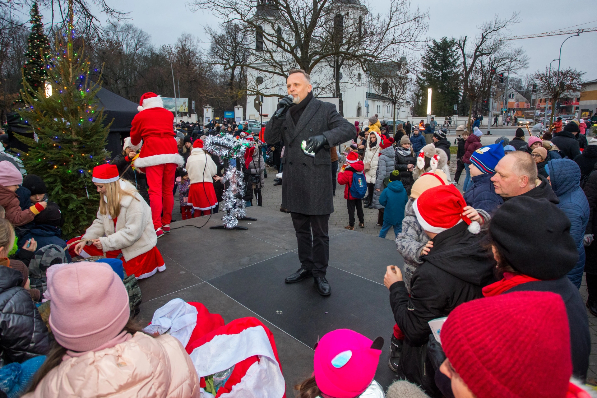 Mężczyzna w ciemnym płaszczu z mikrofonem stoi na scenie na zewnątrz. Wokół niego tłum ludzi w ciepłych ubraniach, wiele osób ma czerwone czapki Mikołaja. Po lewej stronie stoi ubrana na czerwono postać Mikołaja przy udekorowanej choince. W tle drzewa bez liści i budynki. Jest dzień, pochmurne niebo.