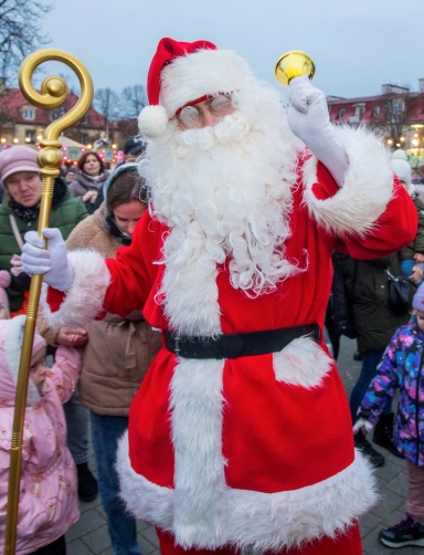 Mężczyzna przebrany za Mikołaja w czerwonym stroju z białym futerkiem, trzyma złoty berło i dzwonek, stoi na zewnątrz wśród ludzi w zimowych ubraniach. W tle budynki i oświetlenie.