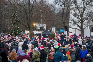 Duża grupa ludzi zgromadzona na zewnątrz w okresie zimowym, wiele osób nosi czapki świąteczne typu Mikołaj, w tle dwa przystrojone choinki i nagłośnienie przy namiocie.