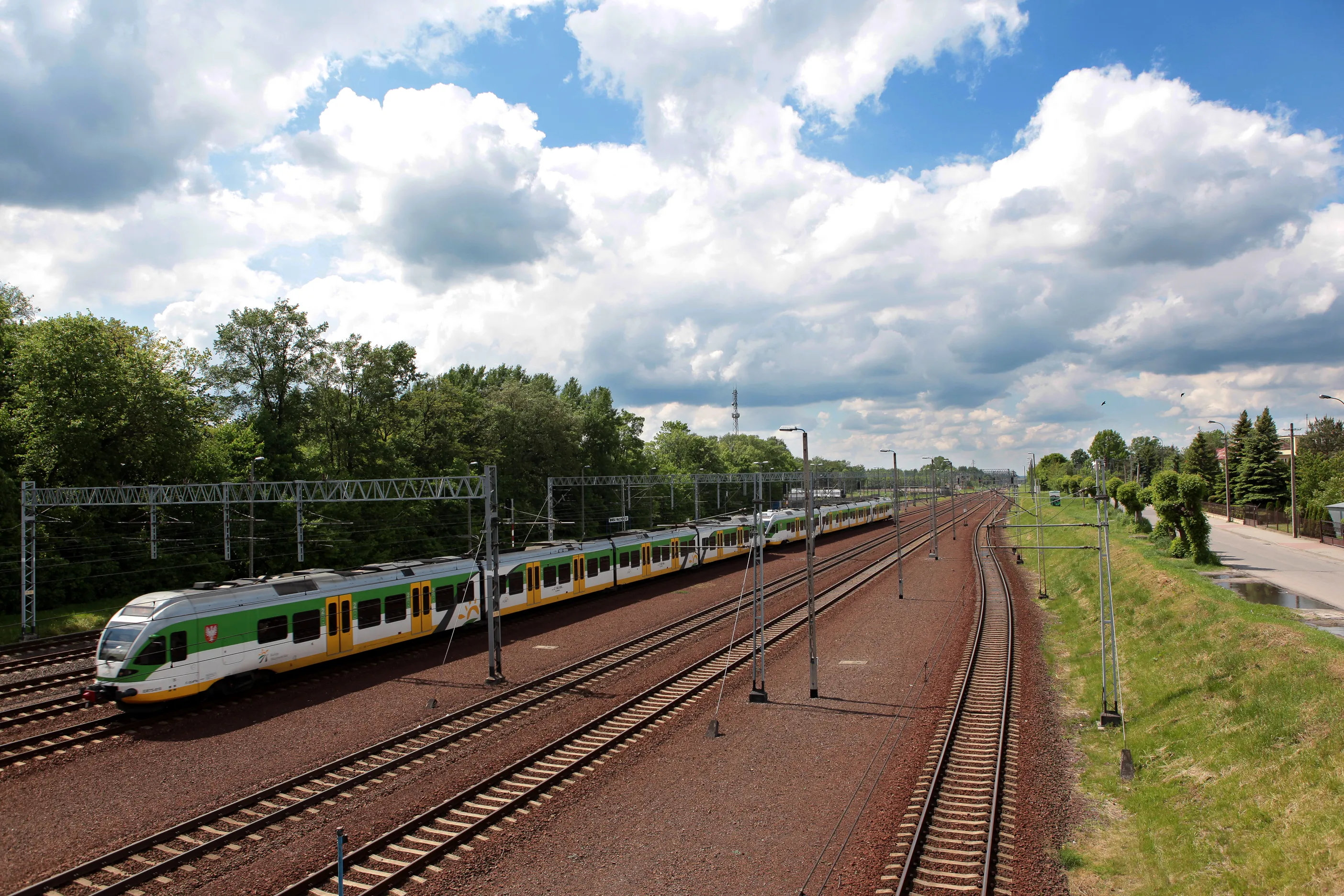 Długi pociąg pasażerski na torach kolejowych obok zielonych drzew pod częściowo zachmurzonym niebem.