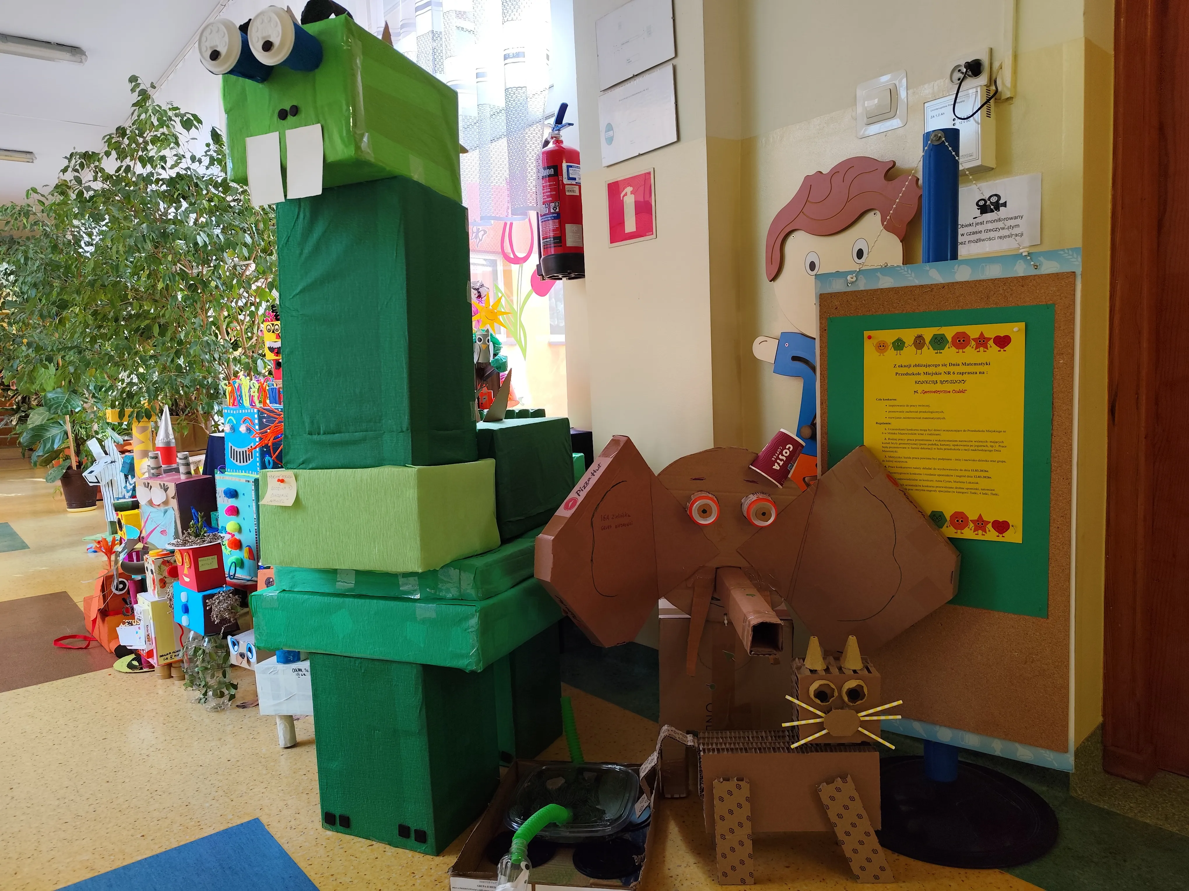 Wewnątrz jasnego korytarza stoi wysoka zielona konstrukcja z pudełek przypominająca krokodyla z białymi zębami i dużymi oczami. Po prawej stoi kartonowy słoń z dużymi uszami. Obok znajduje się mały kartonowy kot z żółtymi wąsami. W tle widać dużą roślinę doniczkową i tablicę ogłoszeń z żółtą kartką.