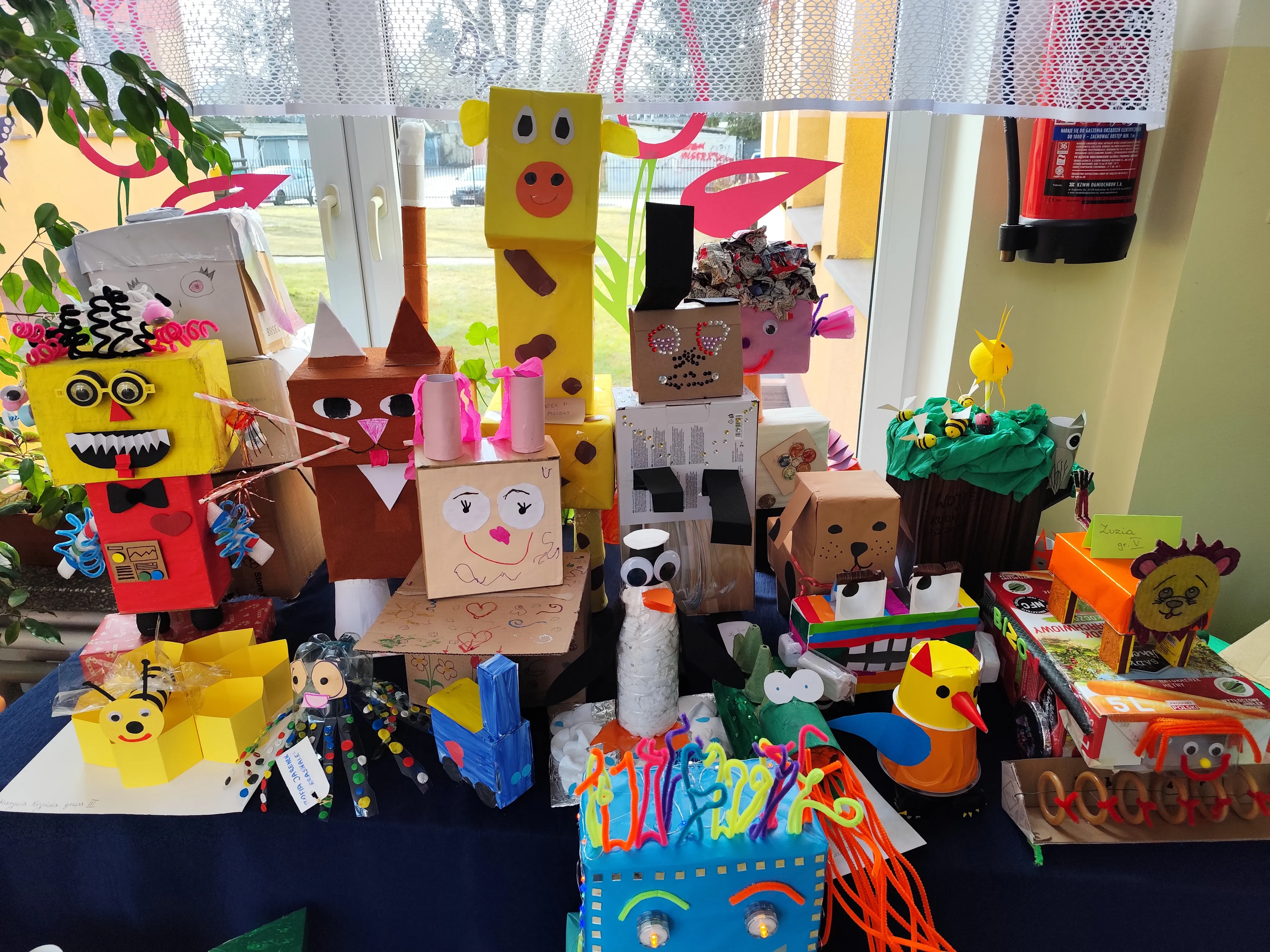 W oknie z białą koronkową firanką stoi stół z wieloma kolorowymi, ręcznie wykonanymi postaciami i pojazdami z pudełek i papieru. Różne figurki mają kształty zwierząt i robotów, dominują żółte i pomarańczowe kolory. Scena jest wewnątrz w ciągu dnia, po prawej widoczny gaśnica na ścianie.