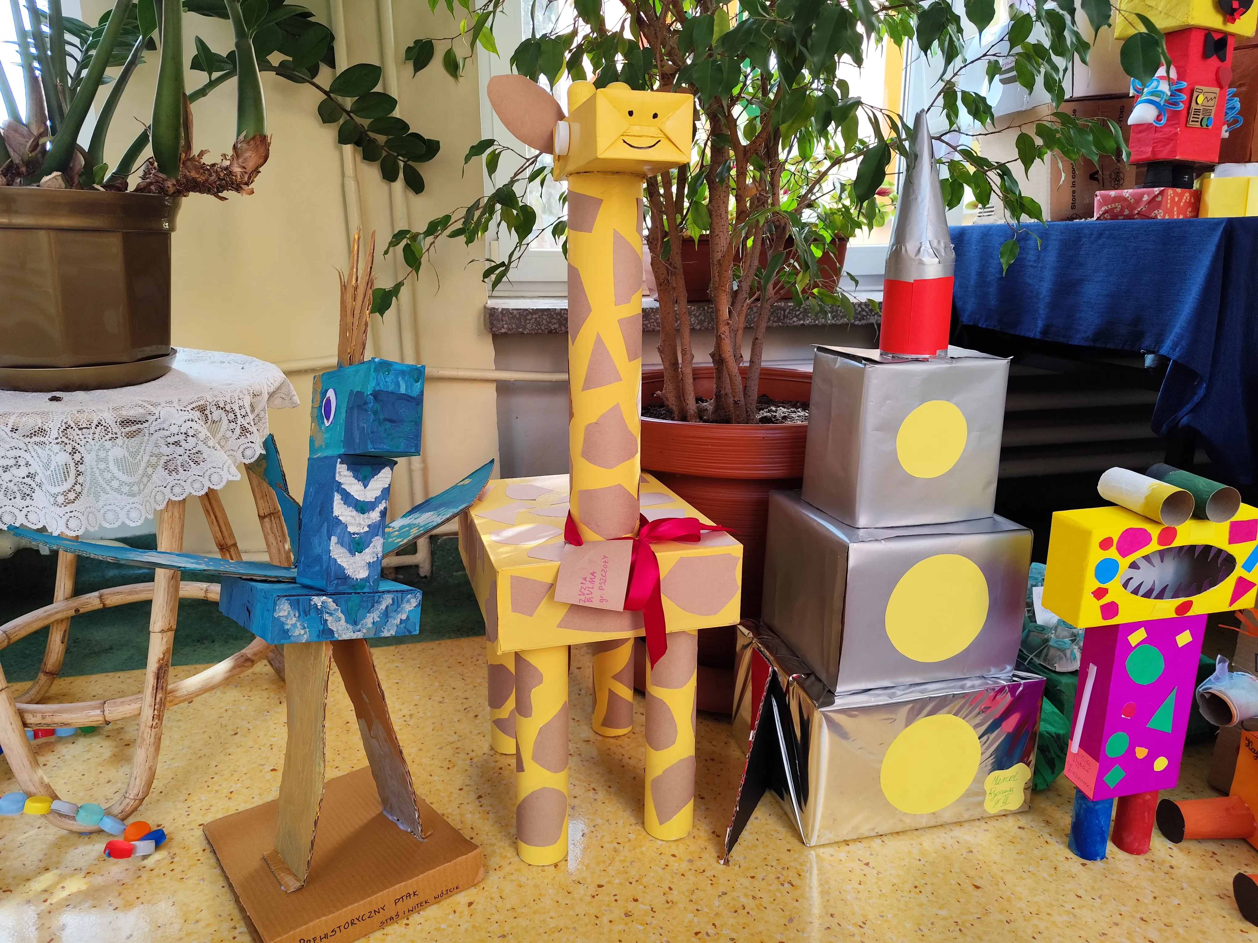 Wnętrze z kilkoma kolorowymi rzeźbami z kartonu ustawionymi na podłodze. Po lewej stoi niebiesa ptakowa konstrukcja na tekturowej podstawie. W środku stoi wysoka żółta żyrafa z brązowymi łatami i czerwoną kokardą. Po prawej są trzy srebrne, nakładające się pudełka z czerwonym stożkiem oraz kolorowe, geometryczne figurki. W tle donica z zielonymi roślinami i stół z koronkowym obrusem.