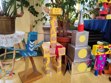 Wnętrze z kilkoma kolorowymi rzeźbami z kartonu ustawionymi na podłodze. Po lewej stoi niebiesa ptakowa konstrukcja na tekturowej podstawie. W środku stoi wysoka żółta żyrafa z brązowymi łatami i czerwoną kokardą. Po prawej są trzy srebrne, nakładające się pudełka z czerwonym stożkiem oraz kolorowe, geometryczne figurki. W tle donica z zielonymi roślinami i stół z koronkowym obrusem.