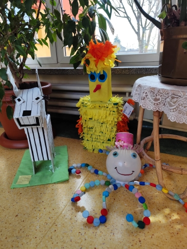 Wewnątrz przy dużym oknie na podłodze stoi trójka papierowych i plastikowych figurek. Po lewej biało-czarna zebra z pionowymi paskami stoi na zielonej podstawie obok doniczki z rośliną. Pośrodku wysoka żółta postać z frędzlowym tułowiem i pomarańczowo-żółtymi włosami. Po prawej uśmiechnięta kula z różowym kapeluszem ma osiem kolorowych odnóży zrobionych z zakrętek. Za figurkami widać parapet, rośliny doniczkowe i mały stolik z koronkowym obrusem.