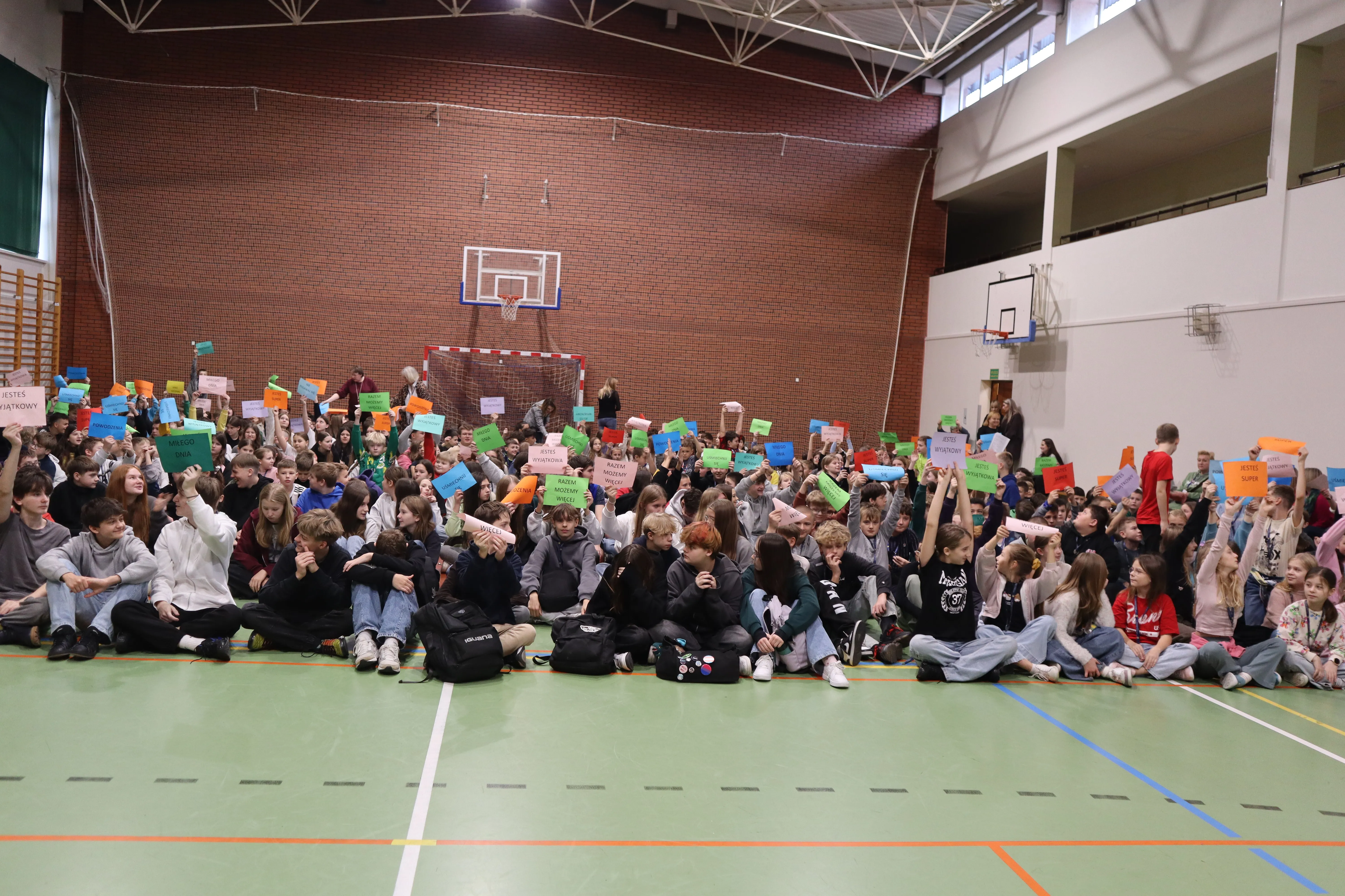 Grupa dzieci i młodzieży siedzi na podłodze hali sportowej. Trzymają kolorowe kartki uniesione do góry. W tle ceglana ściana z bramką do piłki ręcznej i kosz do koszykówki.