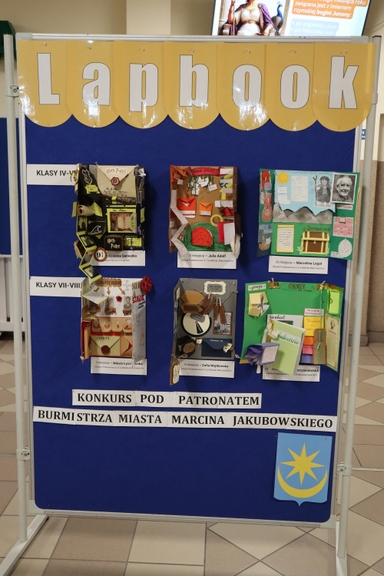 Tablica wystawowa wewnątrz budynku z niebieskim tłem i żółtym banerem z dużymi białymi literami u góry. Na tablicy są trzy rzędowe, kolorowe, przestrzenne lapbooki i projekty szkolne przypięte pinezkami. Na środku i dole znajdują się biało-czarne paski z wydrukowanymi etykietami oraz herb z gwiazdą i półksiężycem po prawej. Płytki podłogowe widoczne pod stojakiem.