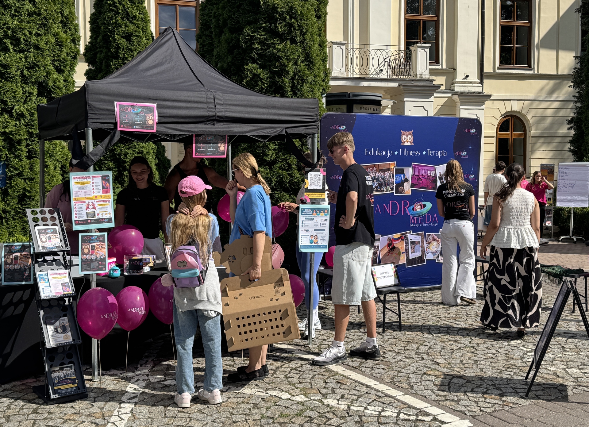 Czarny namiot targowy stoi przed jasnym zabytkowym budynkiem w ciągu dnia. Kilka osób i dzieci rozmawia przy stoisku, jedna osoba nosi różowy plecak. Przy stoisku widać różowe balony, stojaki z ulotkami i duży kolorowy baner informacyjny.