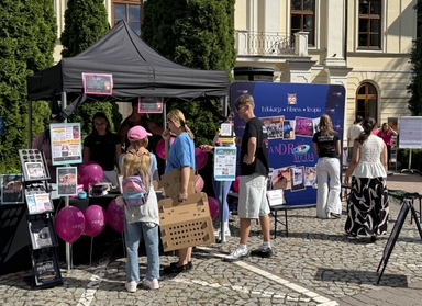 Czarny namiot targowy stoi przed jasnym zabytkowym budynkiem w ciągu dnia. Kilka osób i dzieci rozmawia przy stoisku, jedna osoba nosi różowy plecak. Przy stoisku widać różowe balony, stojaki z ulotkami i duży kolorowy baner informacyjny.