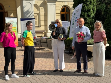 Pięć osób stoi na dziedzińcu przed jasnym kremowym budynkiem w słoneczny dzień. Kobieta w środku przemawia przez megafon, mężczyzna obok trzyma bukiet kwiatów, po bokach widoczne banery i krzewy.