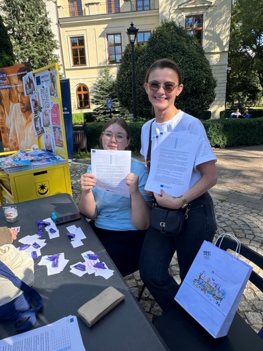 Dwie osoby — kobieta w okularach przeciwsłonecznych i młodsza osoba w okularach — trzymają wydruki z zadaniami i uśmiechają się przy stoliku na zewnątrz w ciągu dnia. Obok stoi żółta tablica promocyjna, wokół rosną krzewy i drzewa, w tle jasny budynek z wieloma oknami. Na stole leżą rozrzucone ulotki i cukierki, obok stoi papierowa torba.