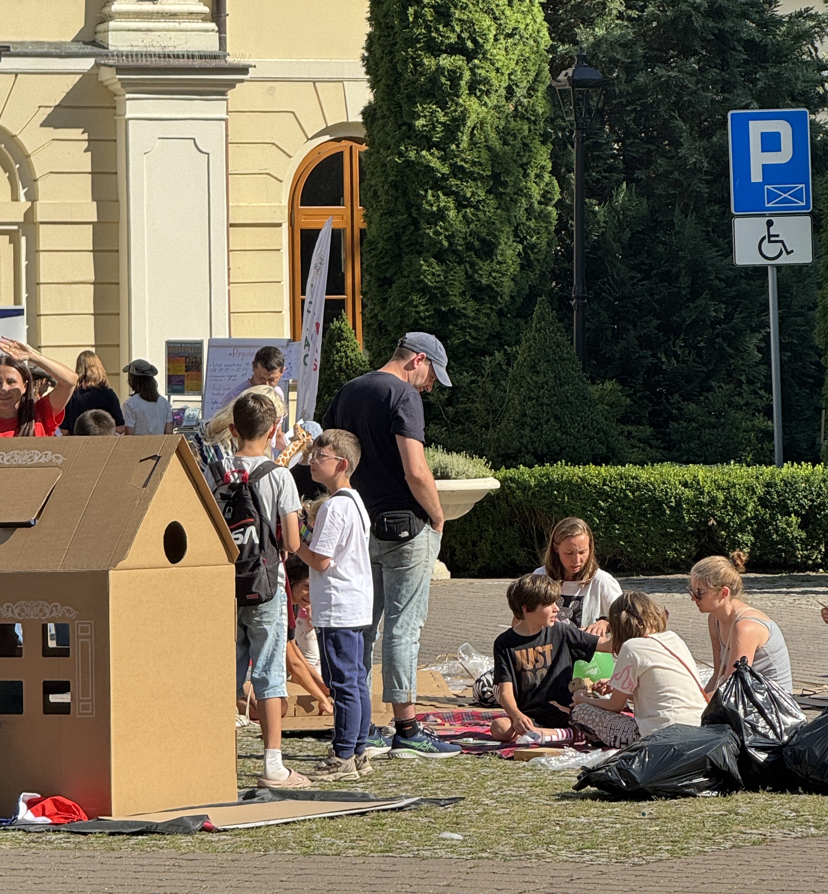 Grupa dzieci i dorośli siedzi i stoi przy dużym kartonowym domku na zewnątrz. Przed nimi leżą koce i zabawki. W tle żółta elewacja z łukowym oknem, wysokie zielone żywopłoty i niebieski znak parkingowy z symbolem wózka inwalidzkiego.