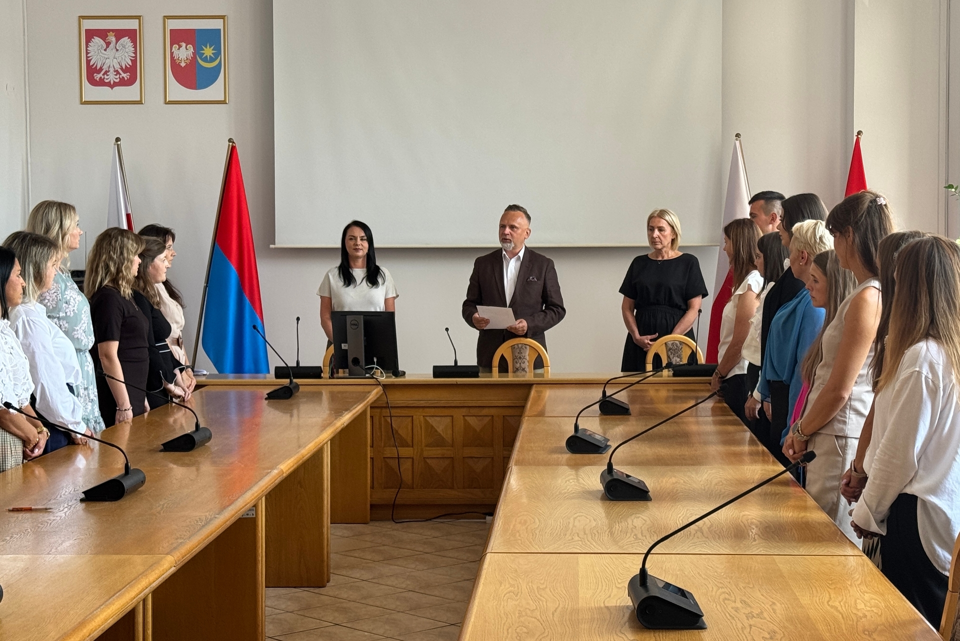 W sali konferencyjnej stoją dwie rzędy ludzi twarzą do środka. Na środku przy stole stoją trzy osoby — mężczyzna z kartką i dwie kobiety. Za nimi wiszą dwa proporczyki i dwie flagi oraz dwa godła na jasnej ścianie. Jasne wnętrze, duży drewniany stół z mikrofonami.