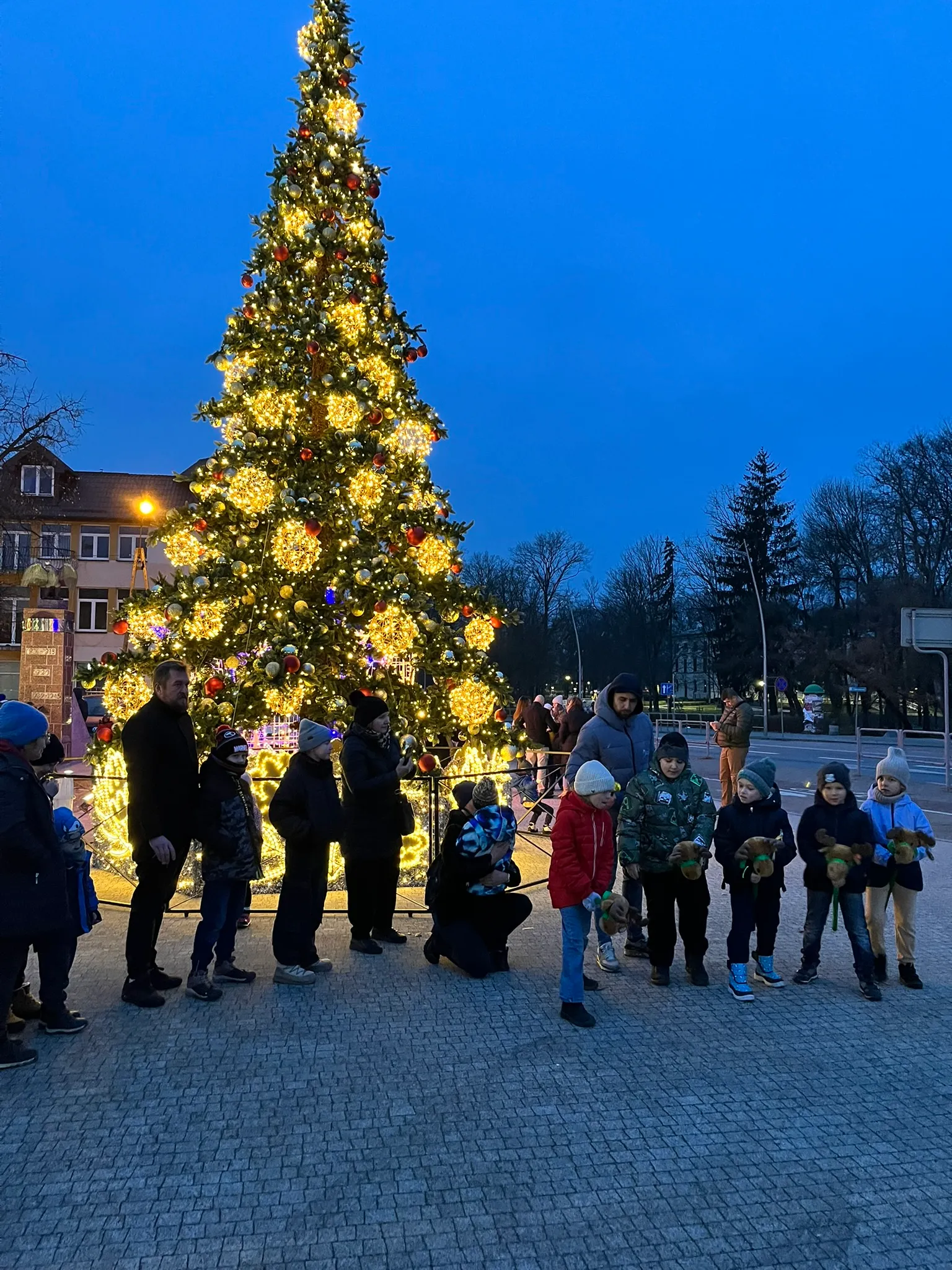 Grupa dzieci i dorosłych stoi na zewnątrz przy wysokiej, ozdobionej świątecznej choince z jasnymi żółtymi i czerwonymi dekoracjami, widoczne wieczorne, niebieskie niebo. Dzieci trzymają pluszowe zabawki. W tle budynki i drzewa bez liści.