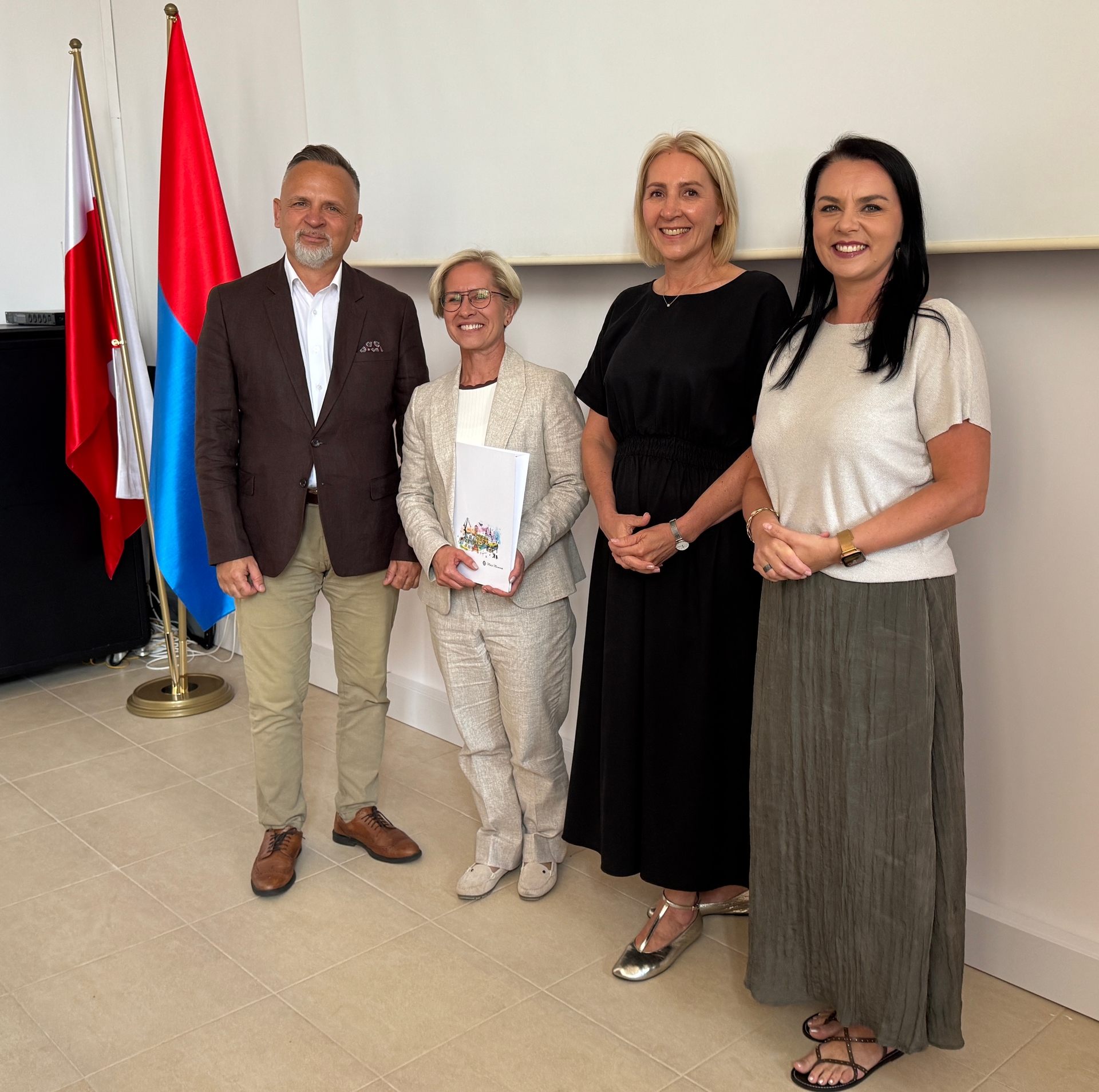 Czterech dorosłych stoi w rzędzie wewnątrz pomieszczenia przed jasną ścianą. Po lewej stoją dwie pionowe flagi — jedna czerwona, druga biało-czerwona. Mężczyzna w brązowej marynarce stoi obok trzech kobiet w jasnej marynarce, czarnej sukience i jasnej bluzce.