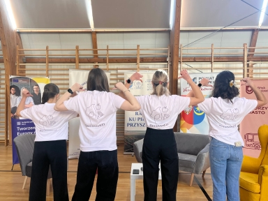 Cztery osoby stoją tyłem w sali gimnastycznej. Osoby ubrano w białe koszulki z prostym logo na plecach. Wszystkie unoszą zgięte ramiona, pokazując obrazek na koszulkach. W tle drewniane drabinki i stojące banery.