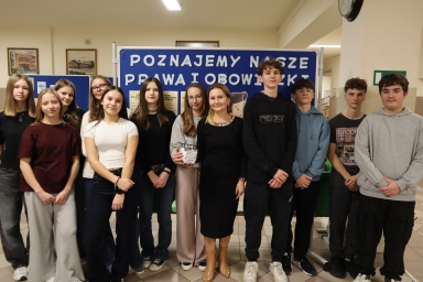 Grupa jedenastu uczniów i nauczycielka stoi w wnętrzu szkoły na tle niebieskiej tablicy z dużym białym napisem. W tle wiszą obrazy, po prawej stronie zegar ścienny. Podłoga jest jasna, pomieszczenie dobrze oświetlone. Uczniowie ubrani w casualowe ubrania.