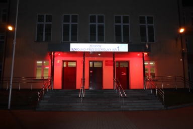 Wejście do budynku szkoły z trzema drzwiami i schodami prowadzącymi do nich, oświetlone na czerwono, z jasną tablicą informacyjną nad drzwiami, widoczne dwa lampy uliczne po bokach.