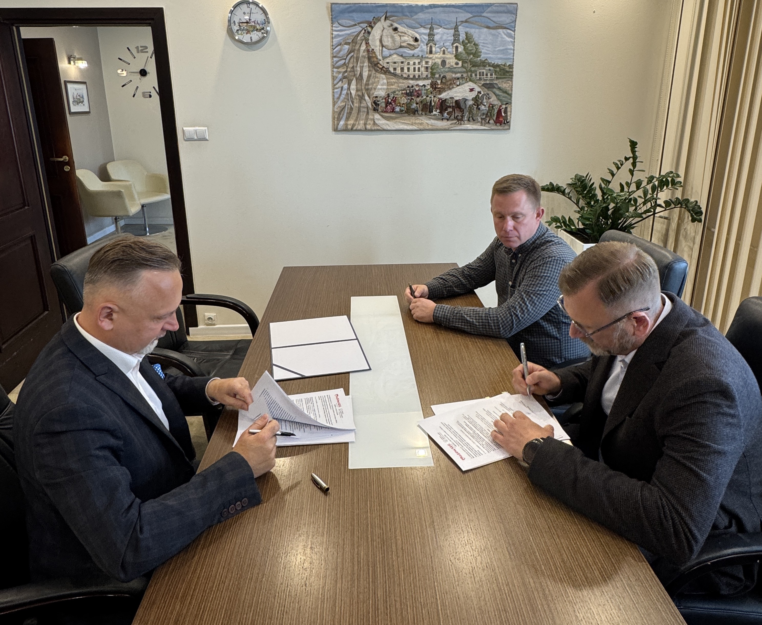 Trzech mężczyzn siedzi przy długim drewnianym stole w biurze i przegląda oraz podpisuje dokumenty. Na ścianie w tle wisi obraz z koniem i budynkami, po prawej stoi duża zielona roślina, po lewej widać otwarte drzwi i zegar.