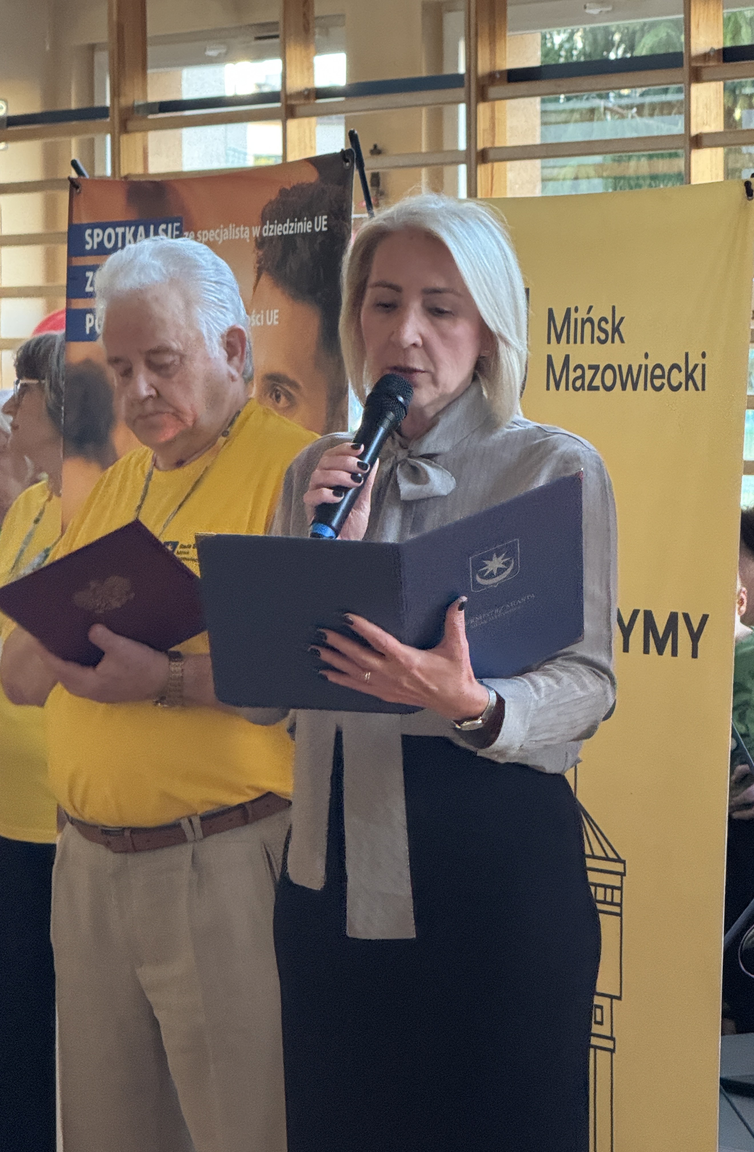 Trzy osoby stoją wewnątrz. Kobieta z jasnymi włosami trzyma mikrofon i czyta z teczki na pierwszym planie. Po lewej stoi starszy mężczyzna w żółtej koszulce trzymający dokument. W tle drewniane okna i pionowy żółty baner z ciemnym napisem.