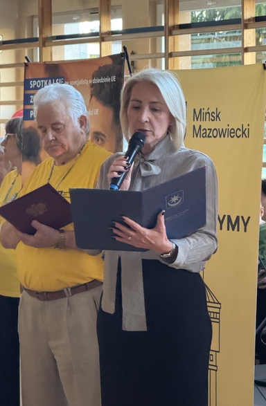 Trzy osoby stoją wewnątrz. Kobieta z jasnymi włosami trzyma mikrofon i czyta z teczki na pierwszym planie. Po lewej stoi starszy mężczyzna w żółtej koszulce trzymający dokument. W tle drewniane okna i pionowy żółty baner z ciemnym napisem.