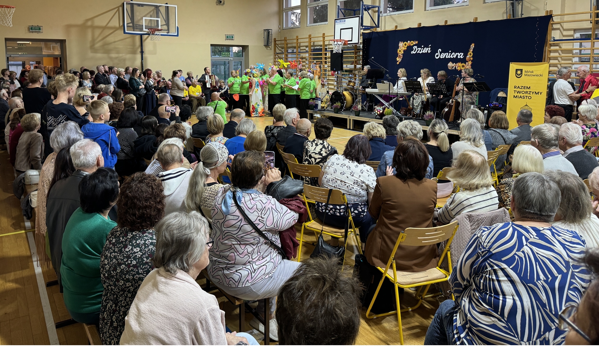 Duża grupa osób starszych siedzi i stoi w sali gimnastycznej podczas wydarzenia. Na scenie z prawej gra zespół muzyczny przy keyboardzie i perkusji. Przed sceną występuje grupa osób w zielonych koszulkach z kolorowymi dekoracjami. Publiczność ogląda i nagrywa telefonami. Sala ma drewnianą podłogę i kosze do koszykówki.