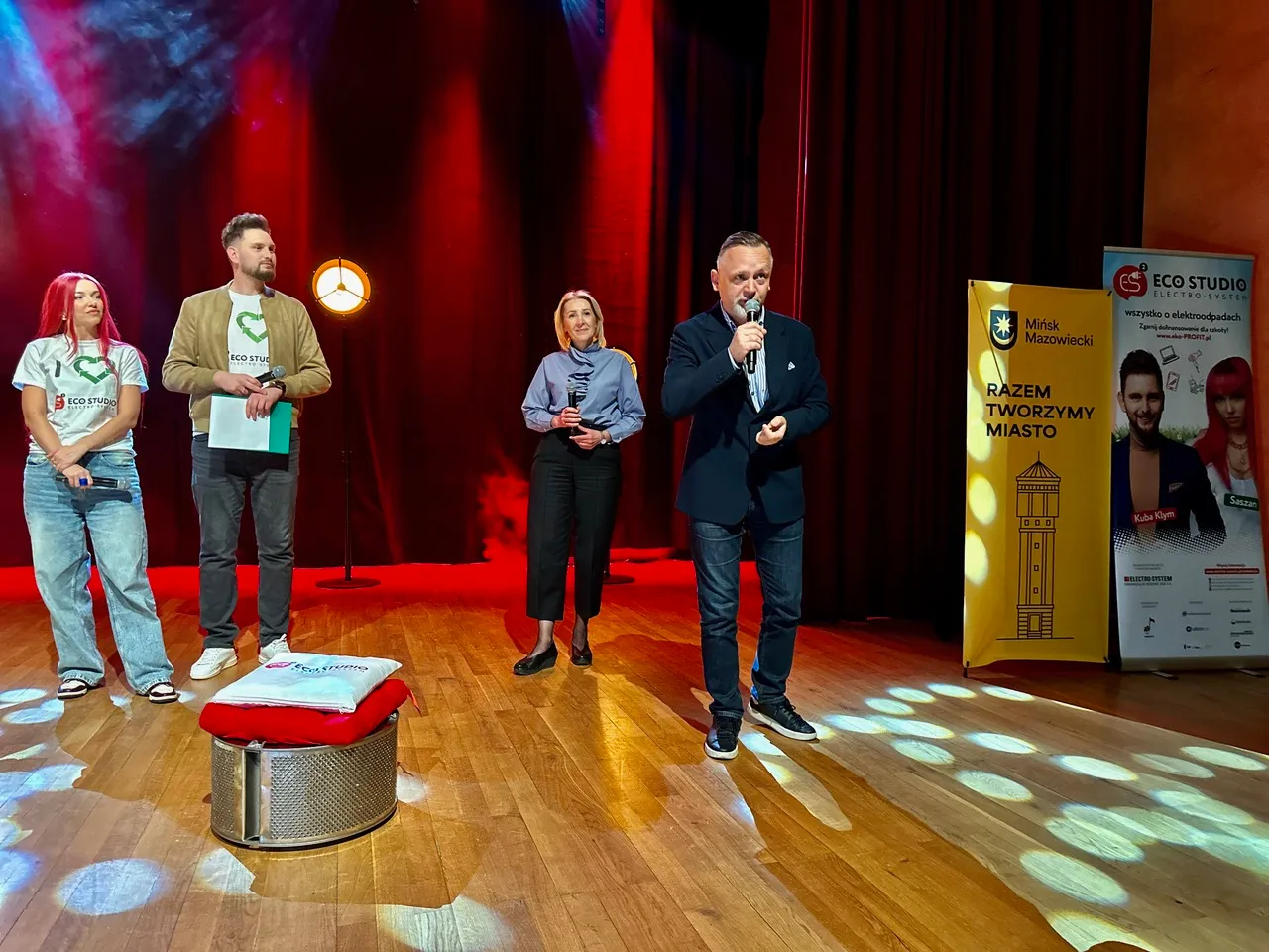 Cztery osoby stoją na drewnianej scenie przed czerwonymi zasłonami; mężczyzna po prawej przemawia do mikrofonu.  
Po lewej dwie osoby w białych koszulkach z zielonym logo i mężczyzna trzymający dokumenty; na środku kobieta trzyma mikrofon.  
Z przodu metalowy bęben z czerwonym podkładem, po prawej dwa stojące banery o żółto-niebieskiej kolorystyce.