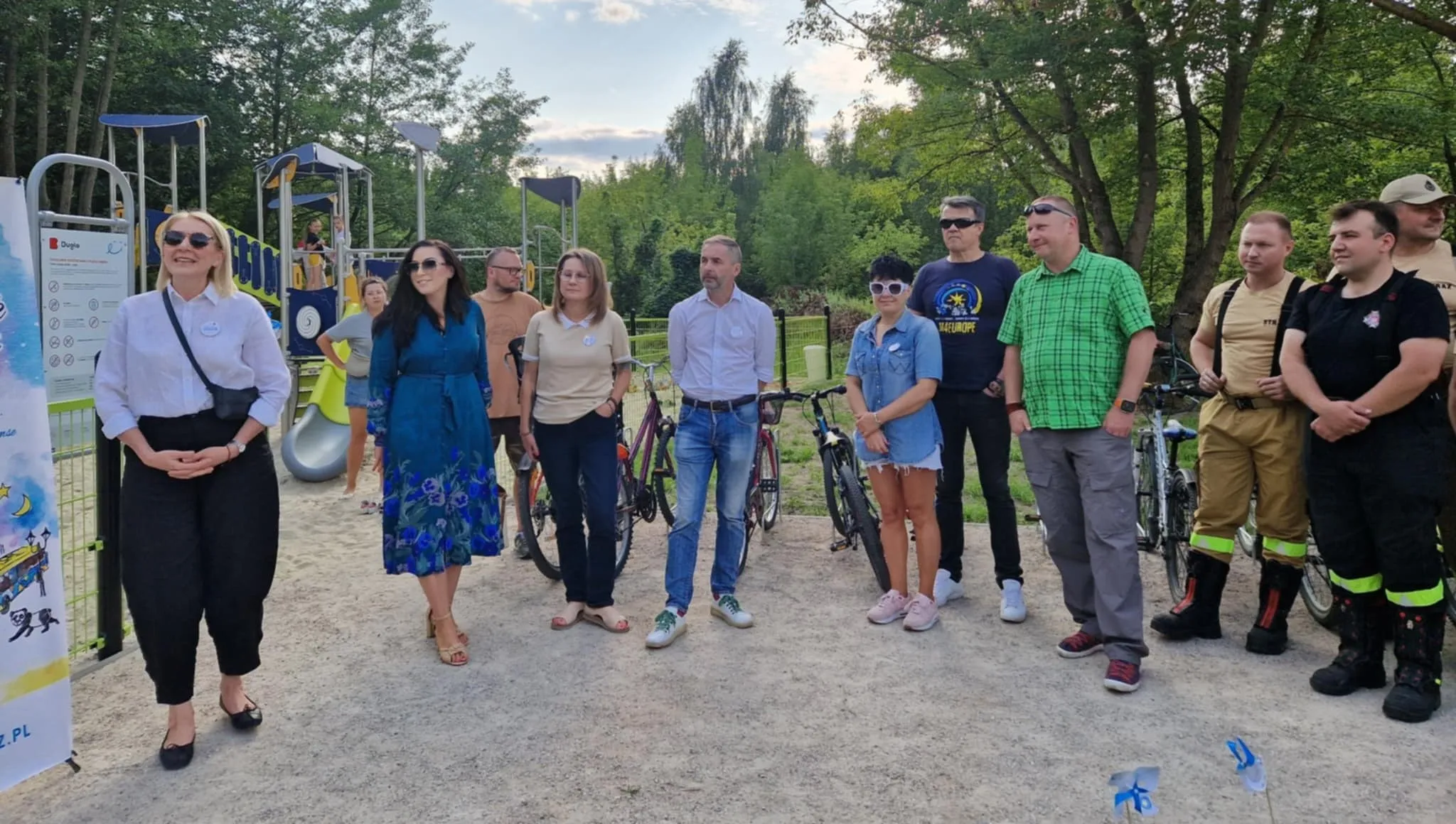 Grupa jedenastu dorosłych osób stoi na zewnątrz na placu zabaw w ciągu dnia, w tle widoczne jest wyposażenie placu oraz zieleń drzew.
