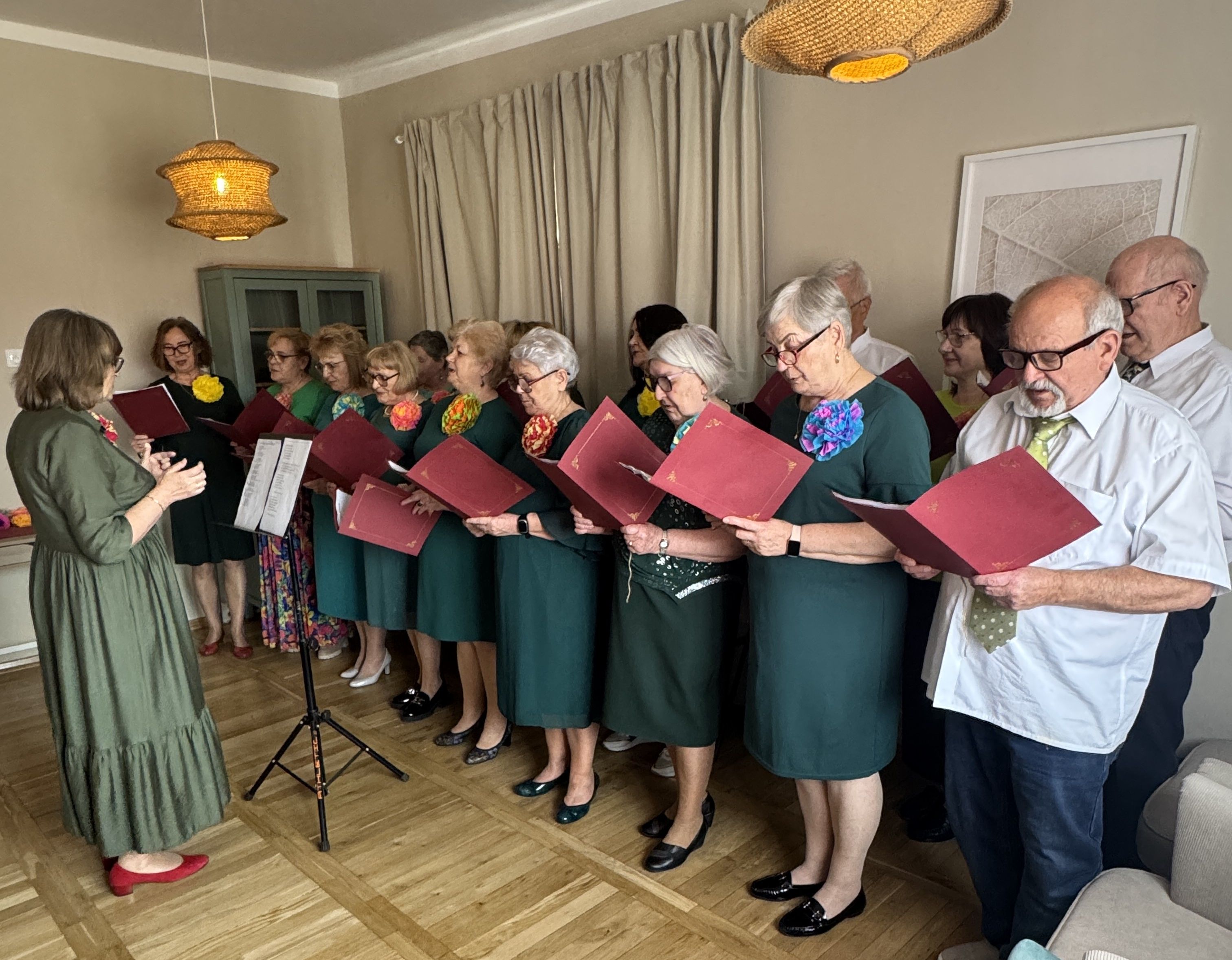 Grupa około piętnastu starszych osób stoi w rzędzie w jasnym pokoju i śpiewa, trzymając czerwone teczki z nutami. Kobiety noszą zielone sukienki z kolorowymi kwiatami przy dekolcie po lewej, dyrygentka w zielonej sukni stoi z przodu po lewej. Dwaj mężczyźni w białych koszulach stoją po prawej, w tle jasne zasłony, szafka i plecione lampy sufitowe.