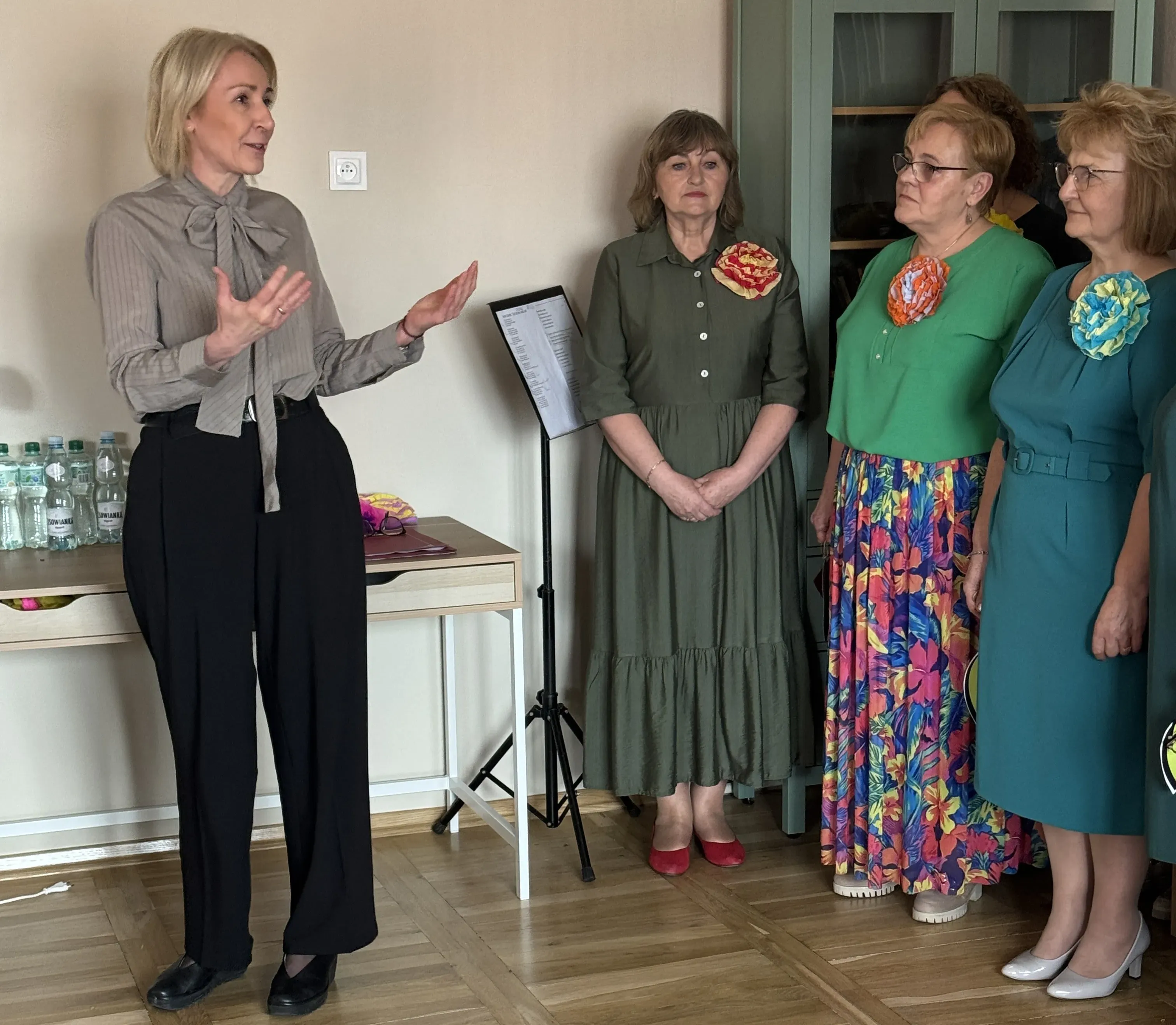 Kobieta po lewej stoi i gestykuluje mówiąc do grupy. Cztery kobiety po prawej stoją obok siebie w kolorowych sukienkach z dużymi kwiatami przy piersiach. Wnętrze z drewnianą podłogą, zieloną szafką, stołem z butelkami wody i nutownikiem na statywie.