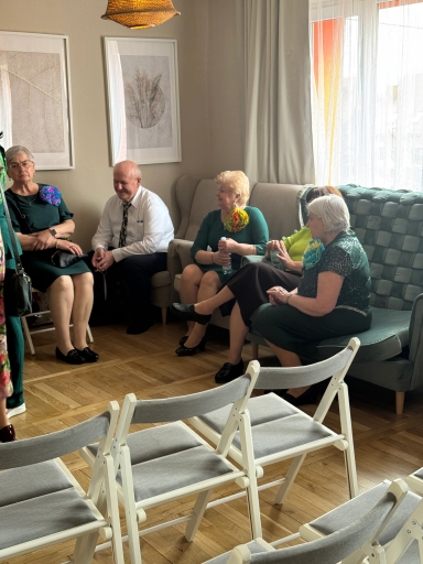 Cztery kobiety i jeden mężczyzna siedzą w salonie przy dużym oknie. Większość osób ma zielone ubrania i kolorowe broszki. Przed nimi ustawione są białe składane krzesła na drewnianej podłodze, na ścianie wiszą dwie ramki z roślinnymi grafikami.