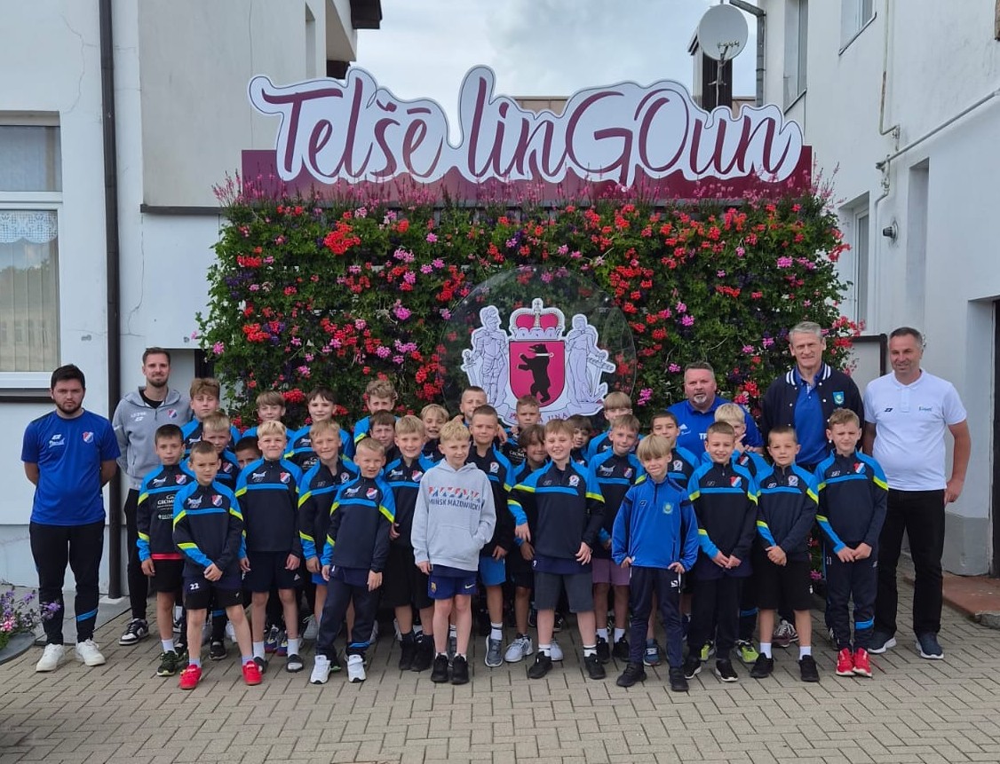 Grupka około trzydziestu chłopców w sportowych strojach stoi na zewnątrz razem z pięcioma dorosłymi trenerami. Za nimi znajduje się pionowa ściana gęsto obsadzona różowo-czerwonymi kwiatami z dekoracyjnym herbem i dużym szyldem u góry, a w tle widać biały budynek i brukowany plac.