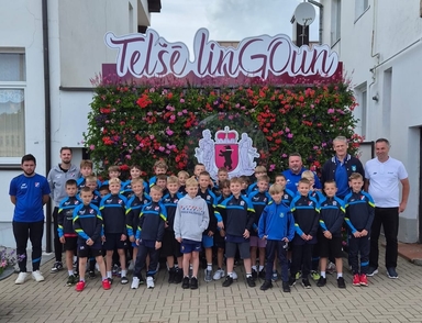 Grupka około trzydziestu chłopców w sportowych strojach stoi na zewnątrz razem z pięcioma dorosłymi trenerami. Za nimi znajduje się pionowa ściana gęsto obsadzona różowo-czerwonymi kwiatami z dekoracyjnym herbem i dużym szyldem u góry, a w tle widać biały budynek i brukowany plac.
