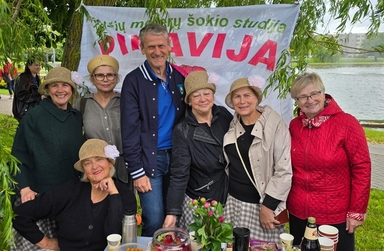 Siedem dorosłych osób — sześć kobiet w słomkowych kapeluszach i jeden mężczyzna — stoi przy stoliku z poczęstunkiem na zewnątrz w dzień pod wierzbą nad brzegiem jeziora. Za grupą wisi baner z dużymi kolorowymi literami, a na stole są kwiaty, kubki i butelki.