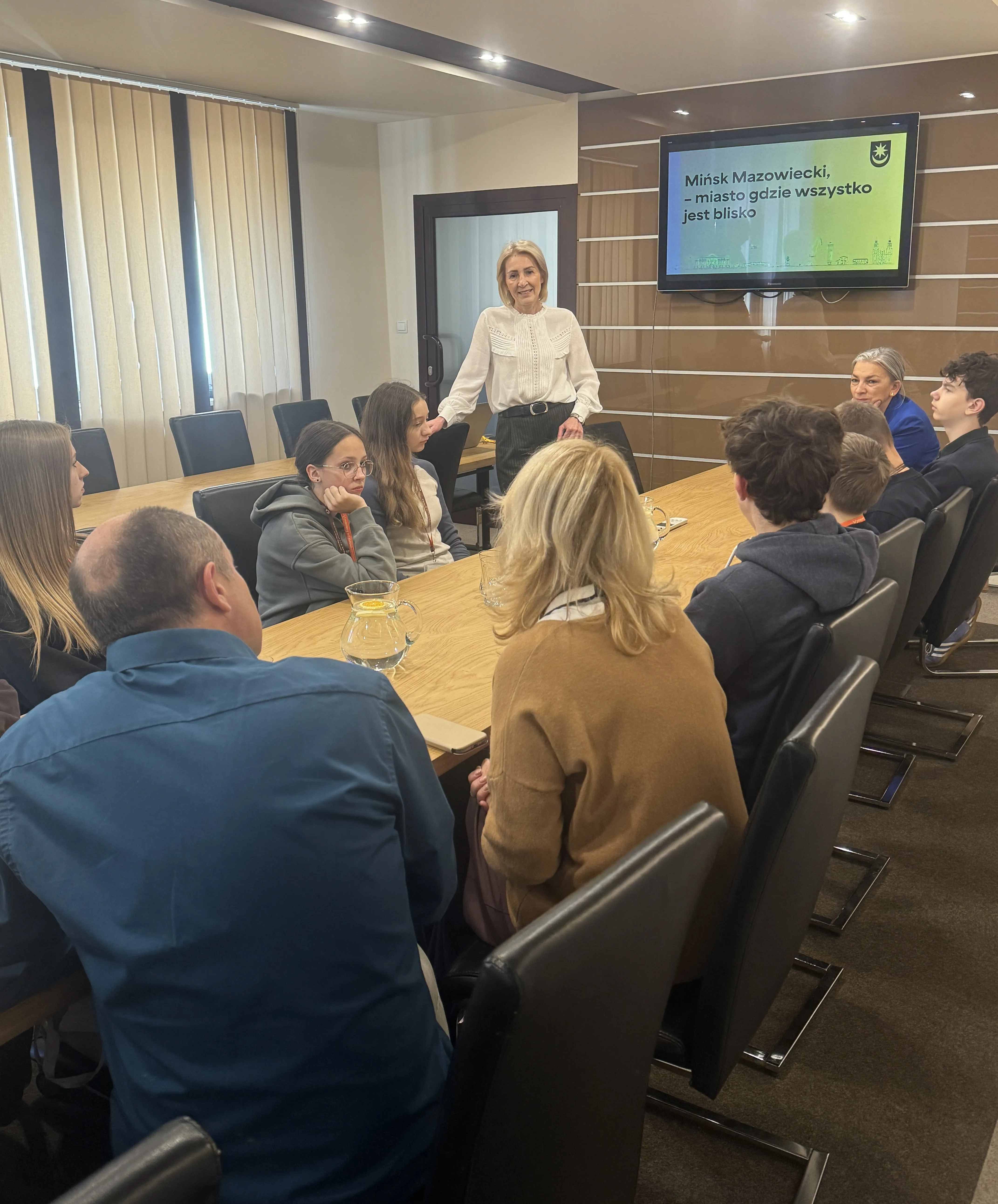 Grupa dziesięciu osób siedzi wokół drewnianego stołu w sali konferencyjnej, jedna kobieta stoi i przemawia. Na ścianie widoczny jest ekran z napisem „Mińsk Mazowiecki, miasto gdzie wszystko jest blisko”. Wnętrze jest jasno oświetlone, okna zasłonięte roletami.