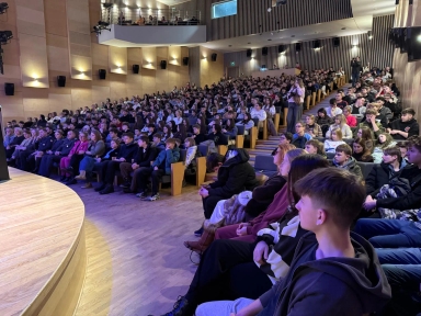 Licząca kilkaset osób publiczność siedzi w amfiteatralnej sali wewnątrz. Rzędy stopniowanych foteli wypełniają prawą i tylną część kadru, po lewej jest zaokrąglona drewniana scena. Jasne drewniane panele ścian i balkon z dodatkowymi widzami są pod ciepłym i fioletowo-niebieskim oświetleniem.