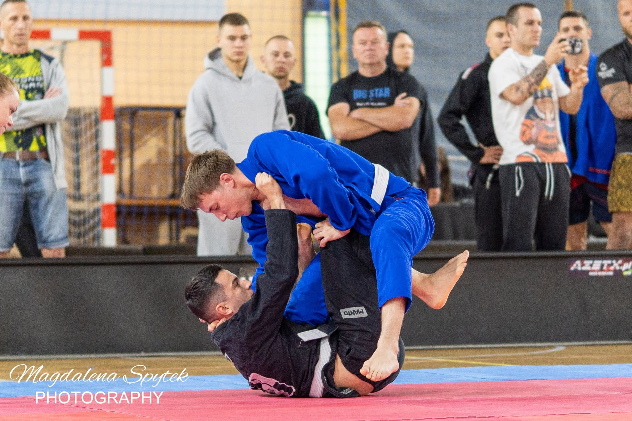 Dwóch zawodników w kimono walczy na czerwono-niebieskiej macie w hali sportowej. Zawodnik w niebieskim kimono jest nad przeciwnikiem, który leży na plecach w czarnym kimonie i obejmuje nogami rywala. W tle stoi grupa kilkunastu widzów obserwujących walkę.