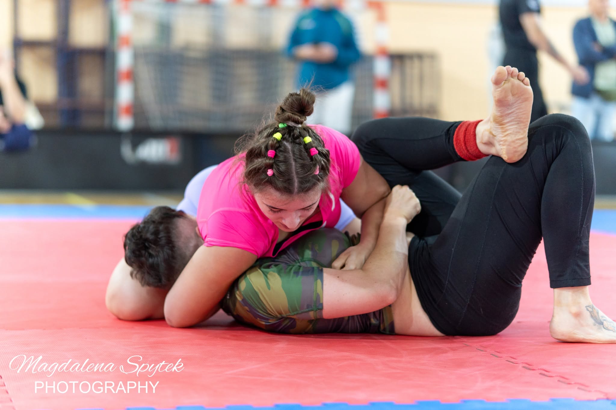 Dwie osoby toczą walkę grapplingową na czerwonym macie w sali sportowej. Kobieta w różowej koszulce z warkoczami i kolorowymi gumkami jest na górze. Druga osoba w czarnych legginsach i moro stroju leży na plecach z uniesioną stopą.