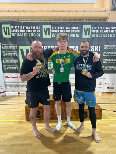 Trzy osoby stoją razem na drewnianym podium w hali sportowej. Młody mężczyzna w zielonej koszulce stoi pośrodku z medalem na szyi. Mężczyzna po lewej ma długą brodę i trzyma medal w ręku, mężczyzna po prawej ma ciemną bluzę i również trzyma medal. Za nimi wiszą sponsorowane banery, na podłodze widać gołe stopy zawodników.