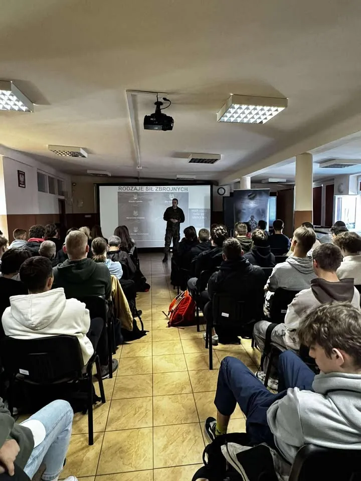 Grupa młodych osób siedzi na krzesłach w sali wykładowej. Mężczyzna w mundurze stoi przed ekranem z rzutnikiem, prowadząc prezentację. W pomieszczeniu jest jasne oświetlenie sufitowe, podłoga jest wyłożona płytkami. Na ekranie widoczny jest tekst dotyczący rodzajów sił zbrojnych.