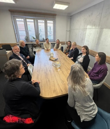 Jedenaście osób siedzi przy półokrągłym drewnianym stole w jasnym pokoju konferencyjnym z dużym oknem i roletami.