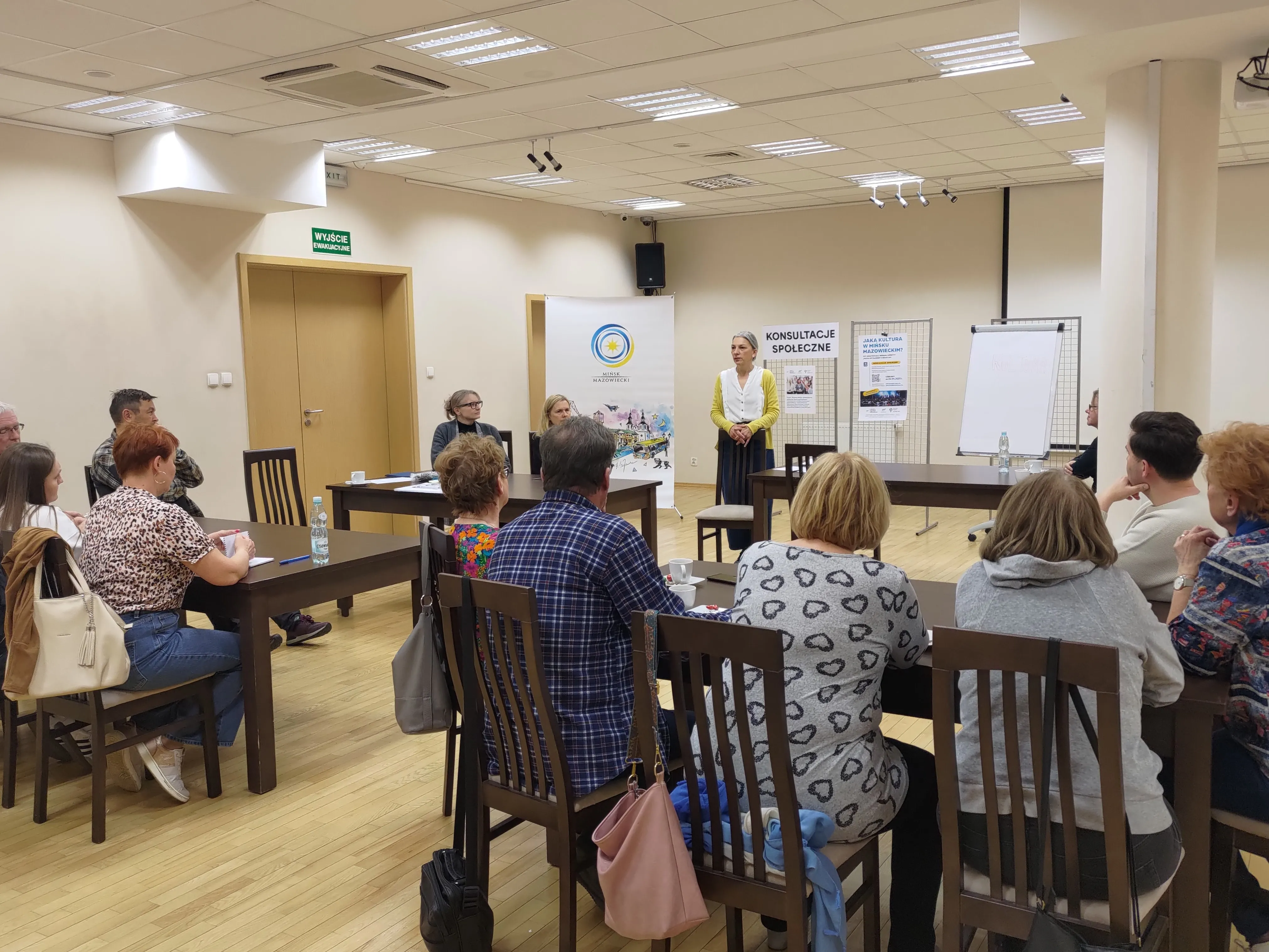 Kilkunastu dorosłych siedzi przy stołach ustawionych w sali konferencyjnej, kobieta na stojąco przemawia do grupy, na ścianie widoczne są plansze z napisami dotyczącymi konsultacji społecznych.