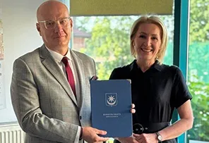 Mężczyzna w szarej marynarce i kobieta w czarnej sukience trzymają dokument z godłem na okładce.