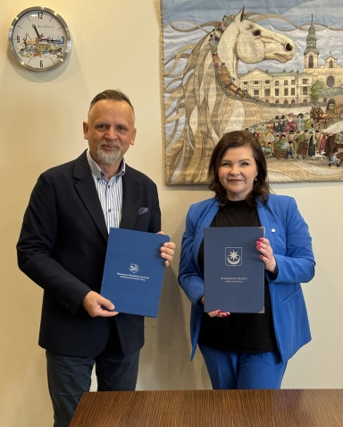 Dwóch dorosłych stoi obok siebie wewnątrz pomieszczenia. Oboje trzymają granatowe teczki i uśmiechają się do aparatu. Na ścianie z tyłu po prawej wisi duża tkanina z wizerunkiem białego konia i sceną miejską, a po lewej widoczny okrągły zegar.