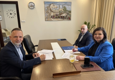 Trzy osoby siedzą przy drewnianym stole w biurze i podpisują dokumenty. Mężczyzna po lewej ma jasną koszulę i granatową marynarkę, mężczyzna na środku siedzi w ciemnym garniturze, kobieta po prawej ma niebieską marynarkę i trzyma długopis. Na stole są teczki, telefon, szklanka z wodą i plasterkiem cytryny oraz rozłożone dokumenty. Na ścianie za nimi wisi gobelin z wizerunkiem konia i zegar; w pomieszczeniu jest jasne, sztuczne oświetlenie.