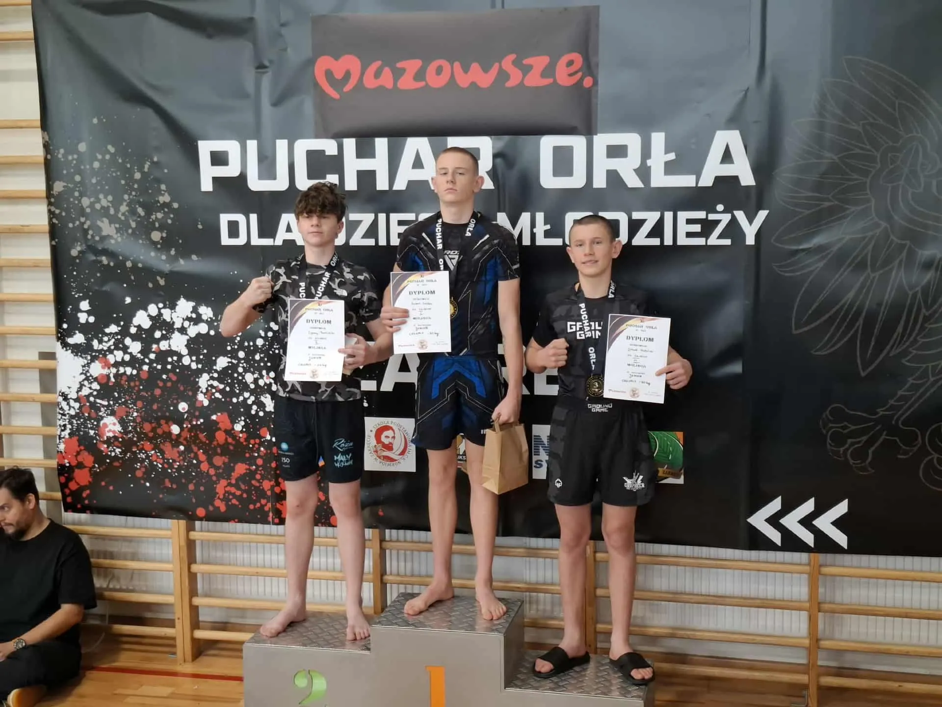 Trzy nastoletnie chłopcy stoją na podium z numerami 1, 2 i 3 w hali sportowej. Każdy trzyma dyplom i ma na szyi medal. W tle czarny baner z napisem „Mazowsze Puchar Orła dla dzieci i młodzieży”. Chłopiec na pierwszym miejscu trzyma także brązową torbę papierową. Na podłodze po lewej stronie siedzi mężczyzna ubrany na czarno.