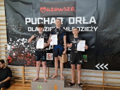 Trzy nastoletnie chłopcy stoją na podium z numerami 1, 2 i 3 w hali sportowej. Każdy trzyma dyplom i ma na szyi medal. W tle czarny baner z napisem „Mazowsze Puchar Orła dla dzieci i młodzieży”. Chłopiec na pierwszym miejscu trzyma także brązową torbę papierową. Na podłodze po lewej stronie siedzi mężczyzna ubrany na czarno.