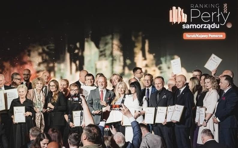Grupa około trzydziestu osób stoi na scenie w rzędach, większość trzyma ramki z certyfikatami. Scena jest wewnątrz, ciemne tło z rozmytymi jasnymi akcentami i logo w prawym górnym rogu. Kilka osób w pierwszym rzędzie robi zdjęcia telefonami, na widowni widać sylwetki i unoszone ręce. Ubrania formalne, dominują ciemne i jasne garnitury oraz sukienki.